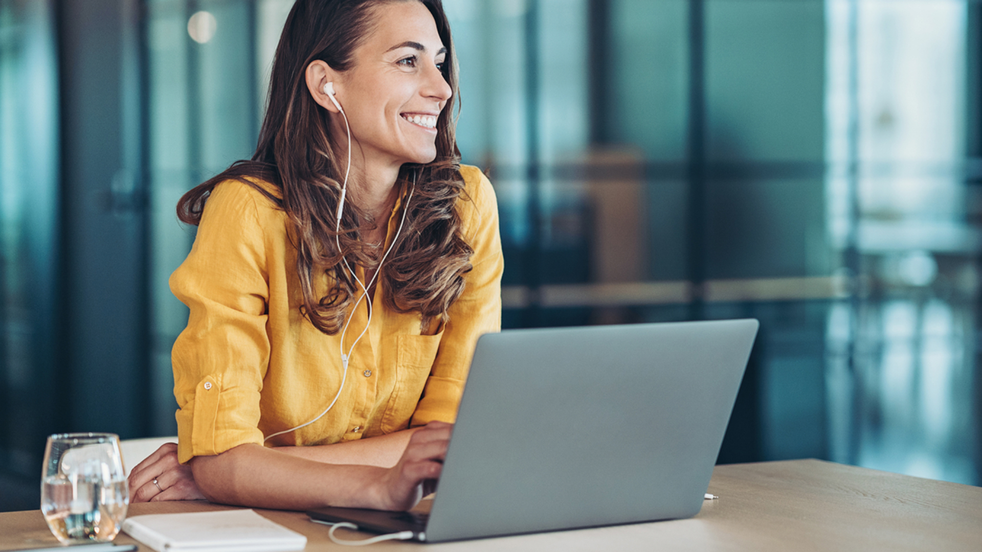 En smilende kvinne med ørepropper sitter foran laptoppen sin og smiler ut i rommet. Foto.