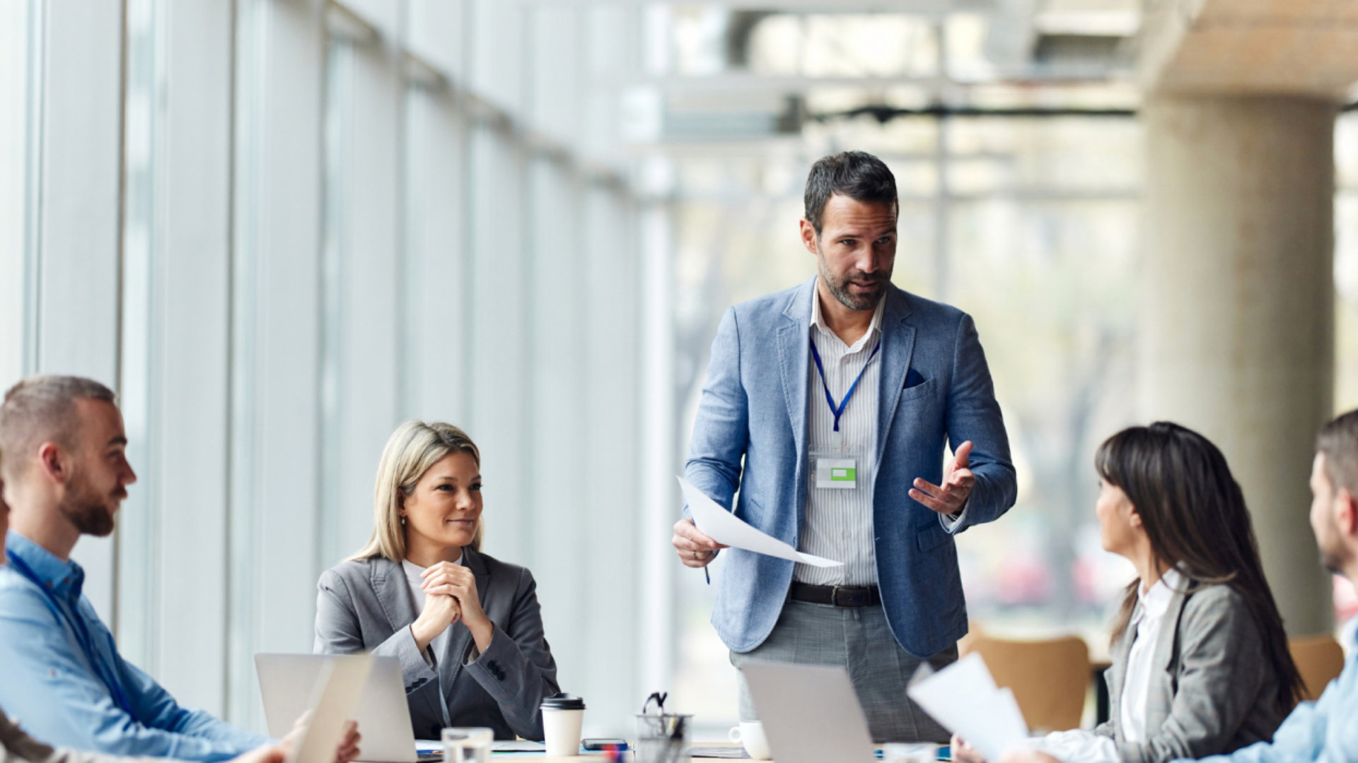 En mann i blå dressjakke står og snakker med flere personer som sitter rundt et møtebord, med laptopper foran seg, også iført dressjakker. Foto.