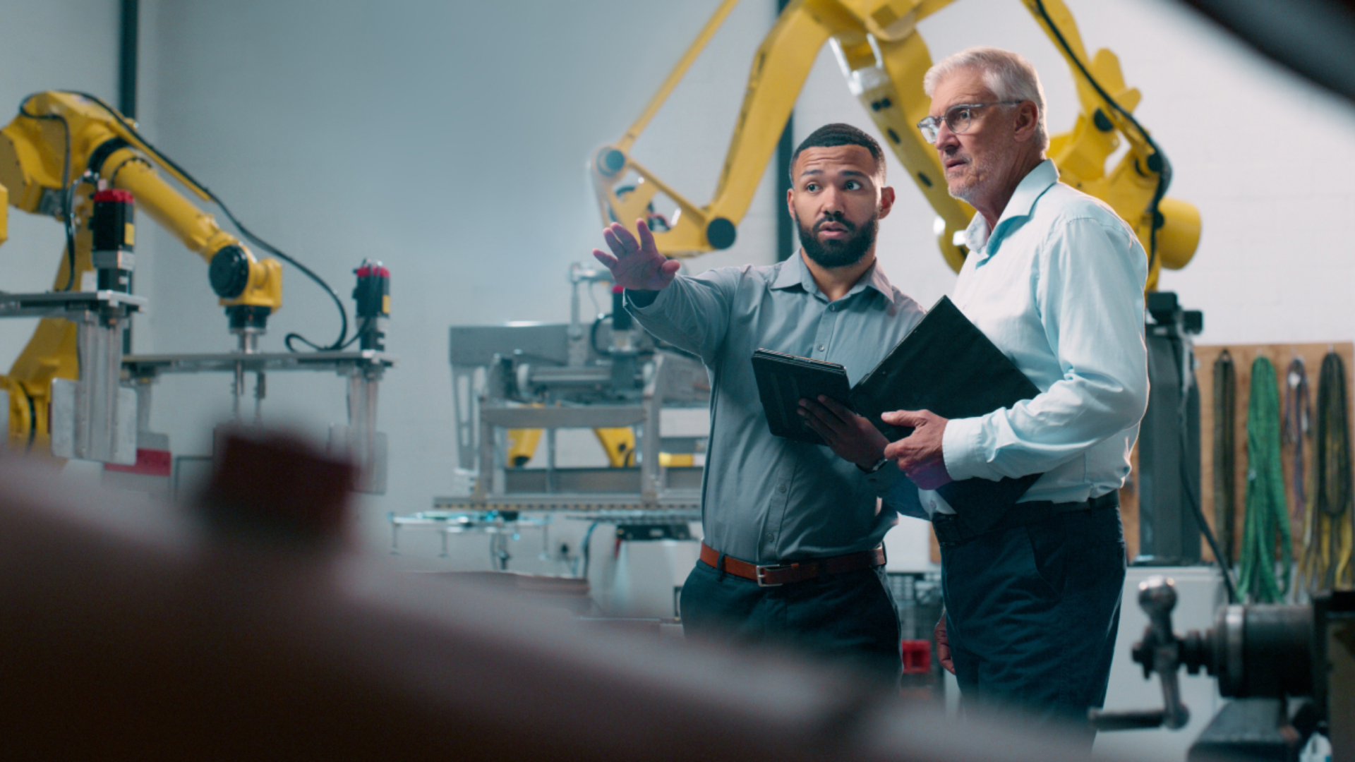 To menn i skjorter står i en fabrikk. Den ene peker utover fabrikken, mens den andre følger blikket i samme retning. I bakgrunnen sees maskiner og en gul kran. Bilde.