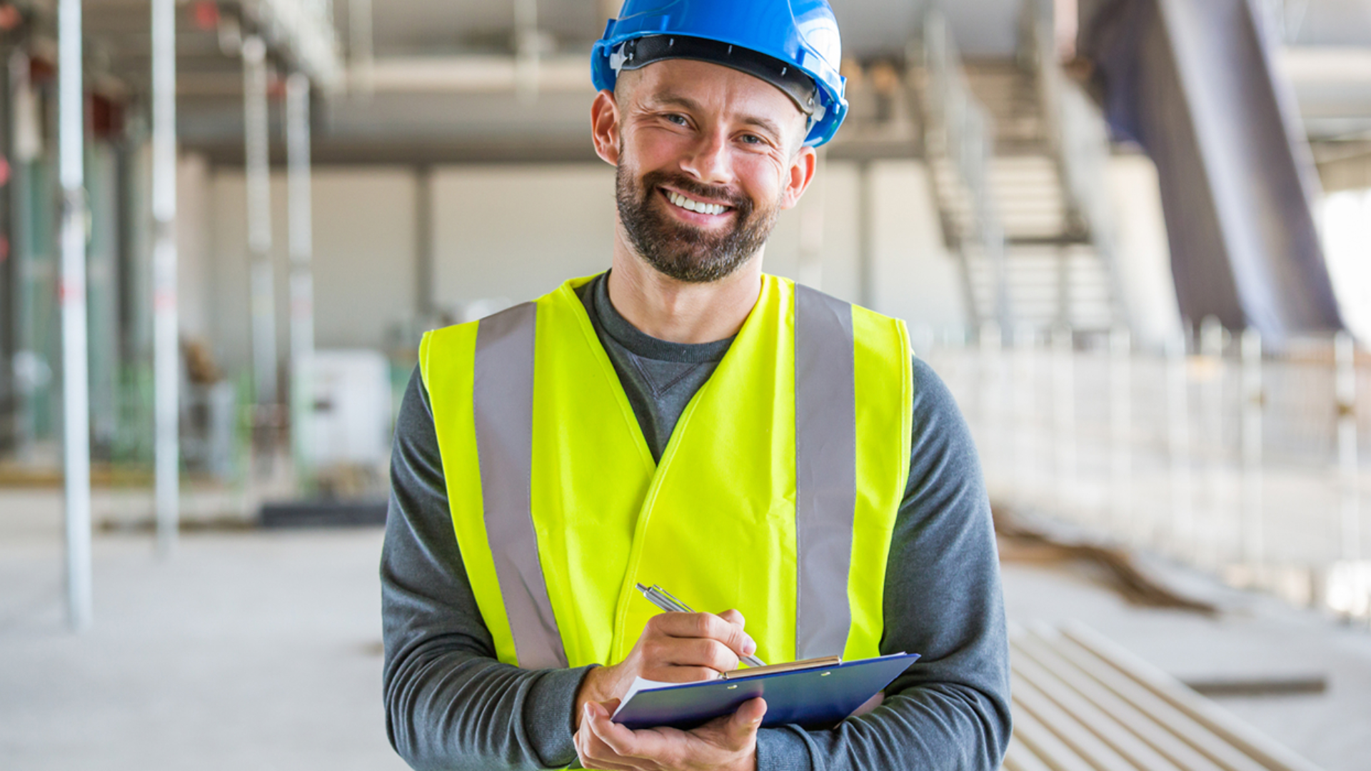 En mann med gul vest og blå vernehjelm smiler. I bakgrunnen sees en byggeplass. Bilde.