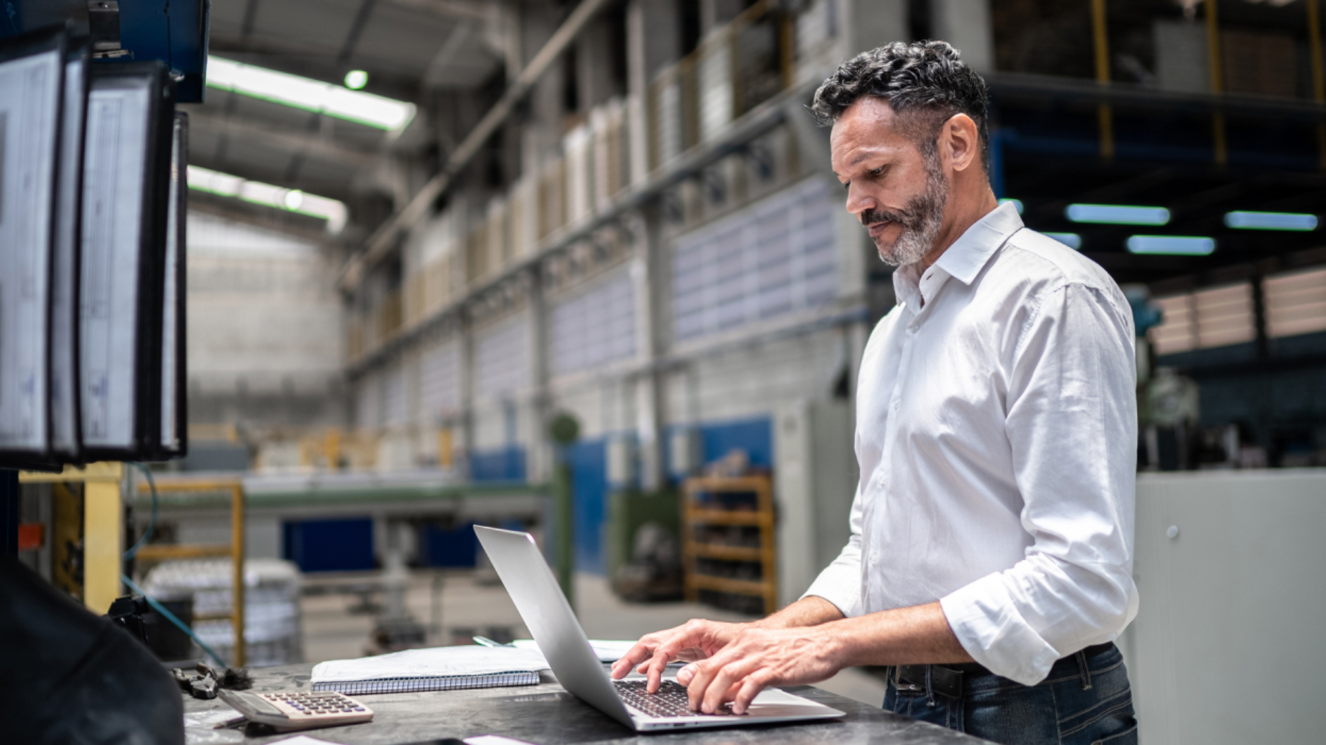 En mann i lys skjorte jobber stående på en laptop. I bakgrunnen sees et industribygg. Bilde.