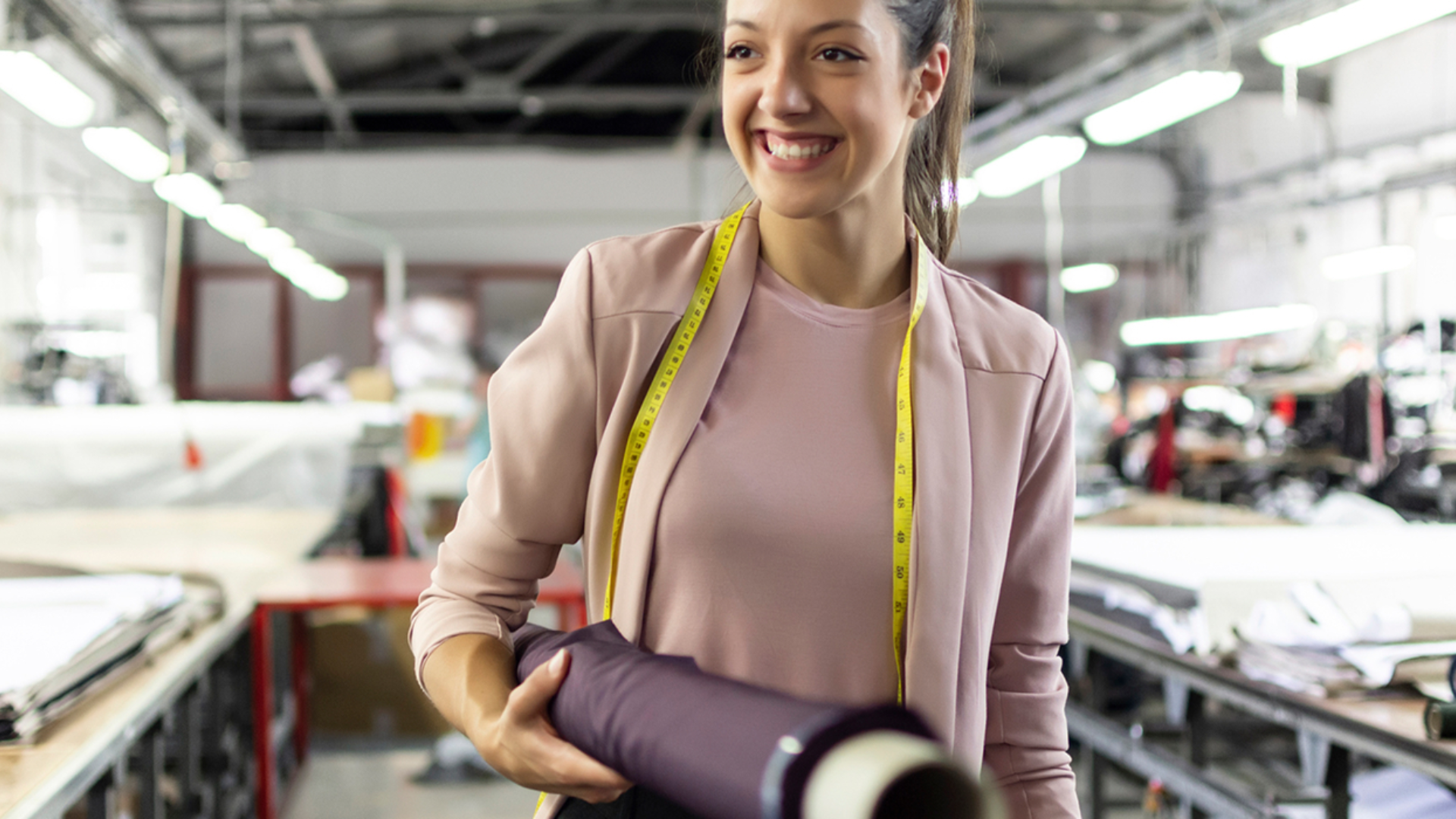 En smilende kvinne med et målebånd rundt nakken og en tekstilrull i hånden, med en tekstilfabrikk i bakgrunnen. Bilde.
