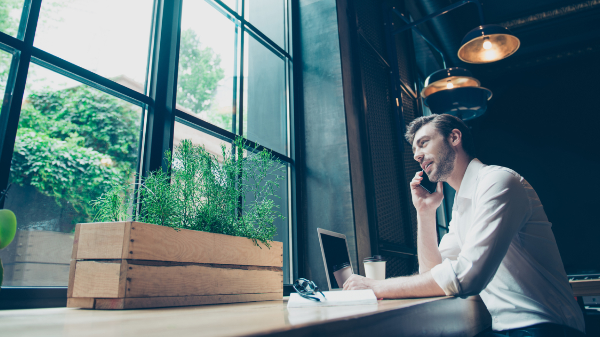 En mann i skjorte sitter med en laptop foran seg, snakker i telefonen og ser ut gjennom et frostet vindu. I bakgrunnen sees et industribygg. Bilde.