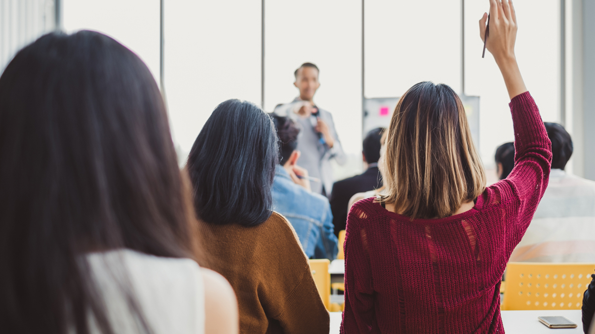"Kvinner og menn sitter med ryggen til, mens en person står foran dem. En kvinne i rød topp har hånden hevet. Foto.