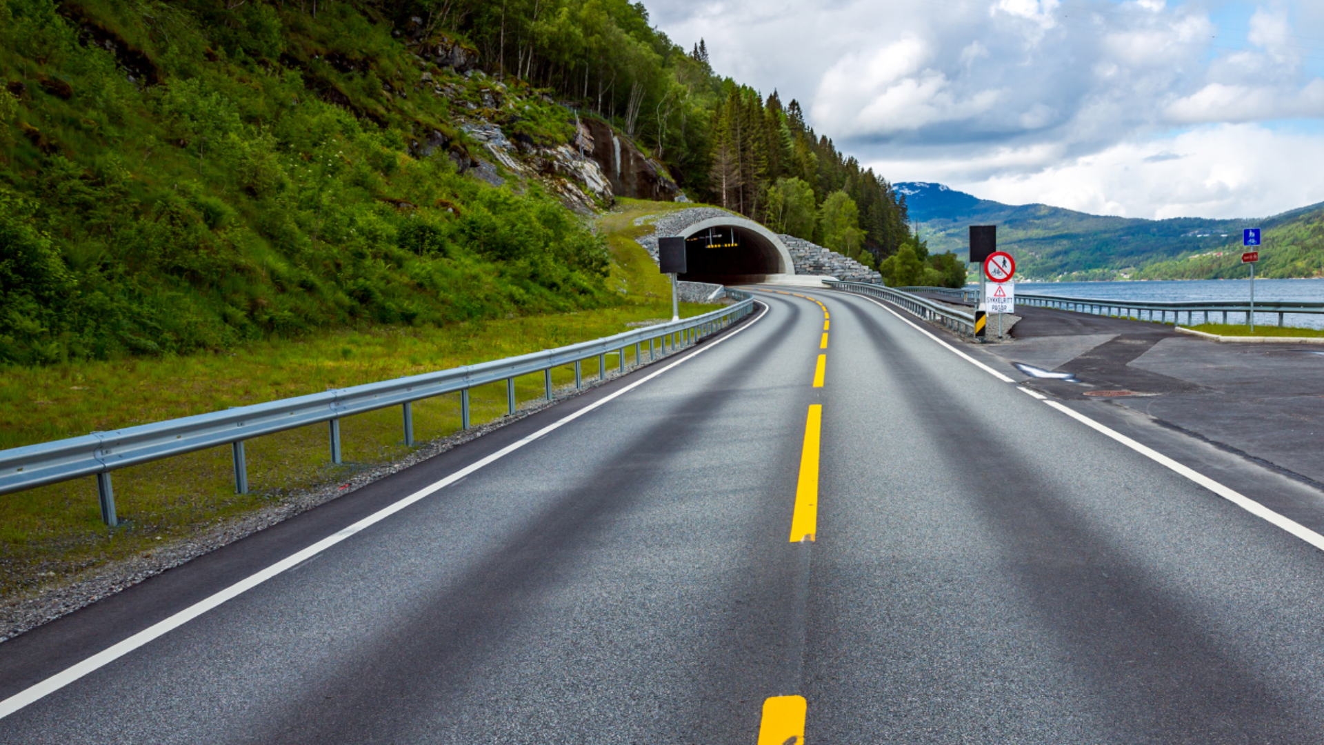 En motorvei leder inn i en tunnel i forgrunnen. Over tunnelen vokser grønne trær, og til høyre for den ligger et vann omgitt av fjellkjeder. Foto.