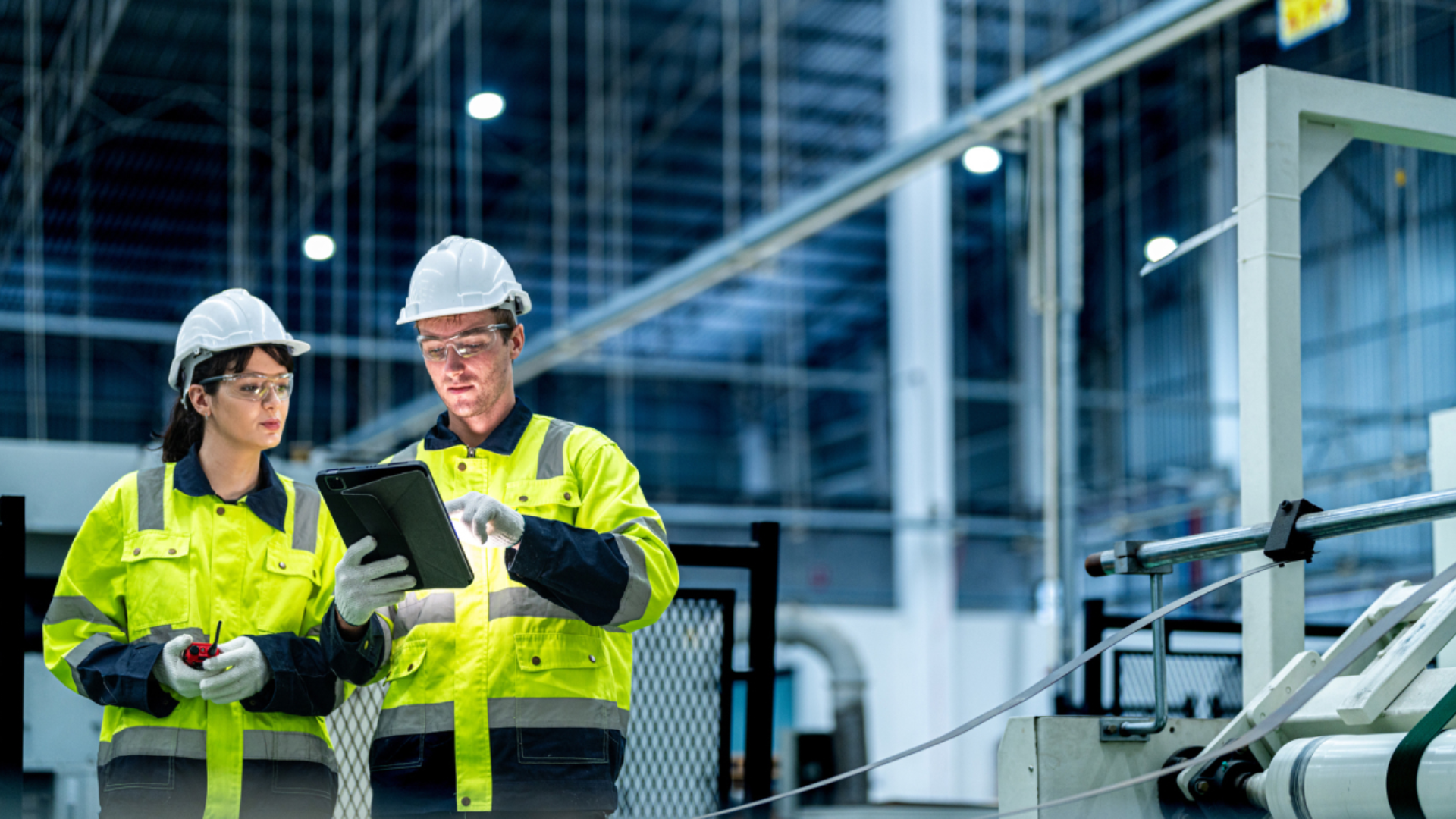 To industriarbeidere iført vernehjelmer, vernebriller og gule refleksjakker står i en moderne fabrikkhall. Den ene holder et nettbrett og peker på skjermen, mens den andre lytter og holder et verktøy. Maskiner og produksjonsutstyr er synlige i bakgrunnen. Foto.