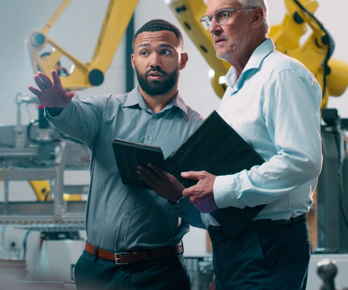 To menn i skjorter står i en fabrikk. Den ene peker utover fabrikken, mens den andre følger blikket i samme retning. I bakgrunnen sees maskiner og en gul kran. Foto.