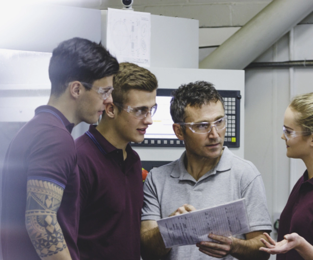 Tre menn med vernebriller står sammen, hvor den ene holder et ark og peker mens de ser på en kvinne, også med vernebriller, og snakker med henne. Foto.