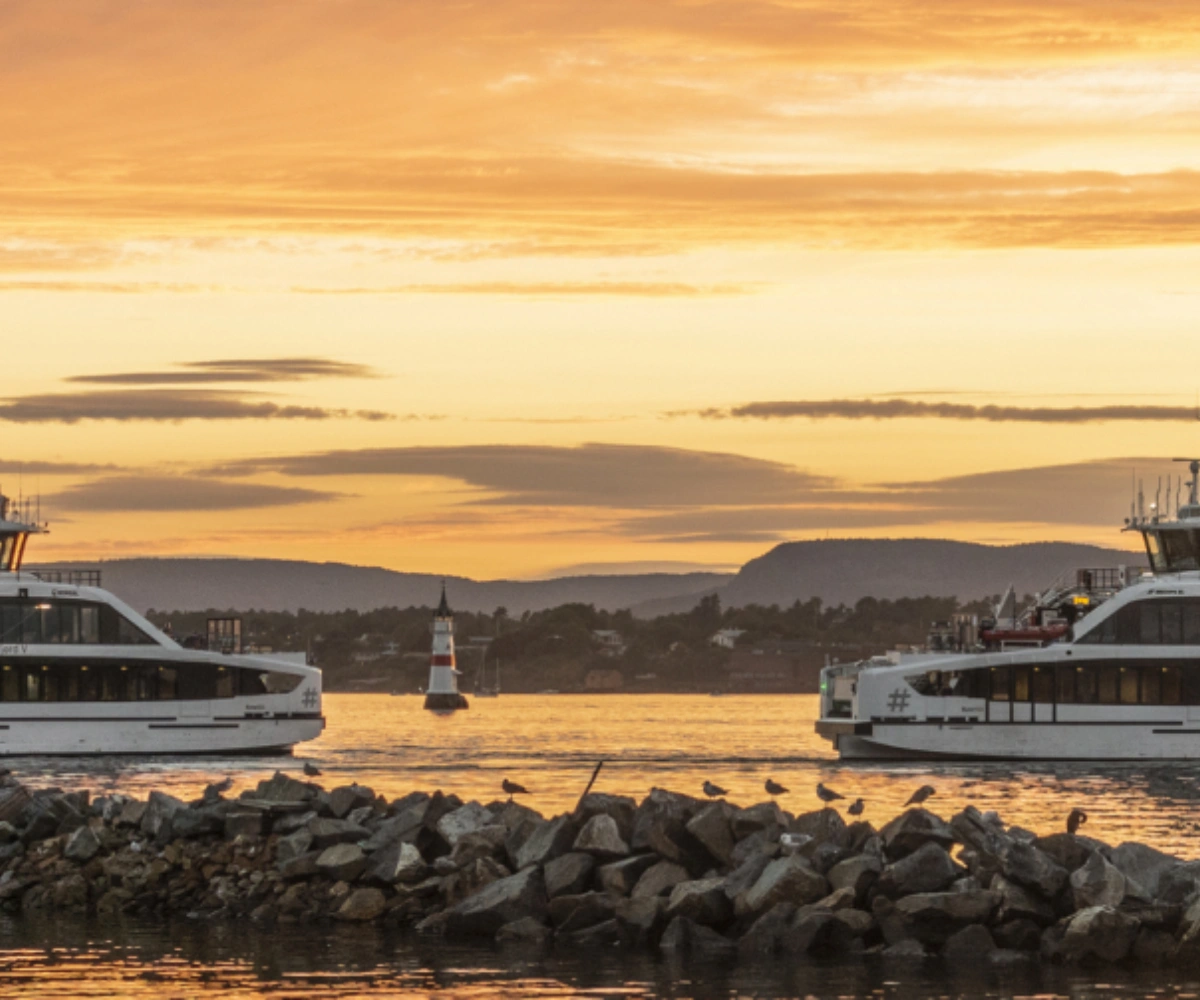To båter kjører over vannet i gyllent kveldslys under golden hour, med en by langt i bakgrunnen. Foto.
