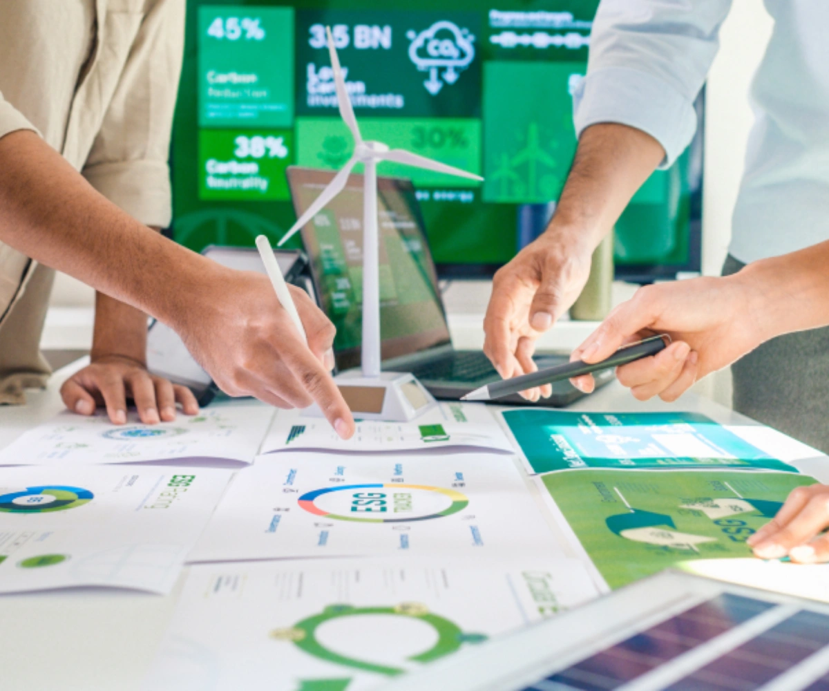 Nærbilde av grafer og illustrasjoner på et bord, med en liten modell av en vindmølle. Rundt bordet er tre personer synlige, hvorav vi bare ser armene deres som holder en penn og et beger over en av grafene. Foto.