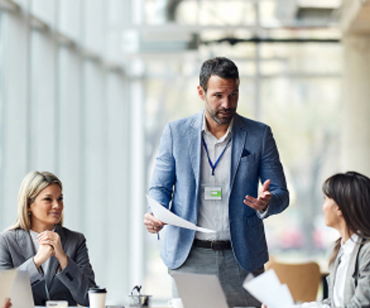 En mann i blå dressjakke står og snakker med to kvinner som sitter med laptopper foran seg, også iført dressjakker. Foto.