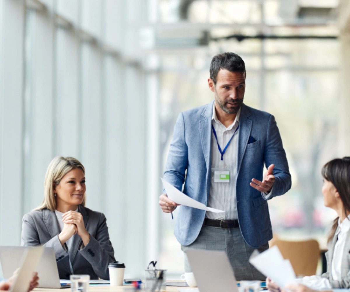 En mann i blå dressjakke står og snakker med to kvinner som sitter med laptopper foran seg, også iført dressjakker. Foto.