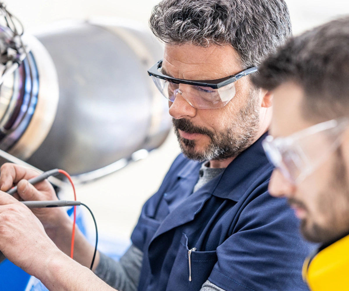 To arbeidere med vernebriller, der den ene holder røde og blå testkabler. Bilde.