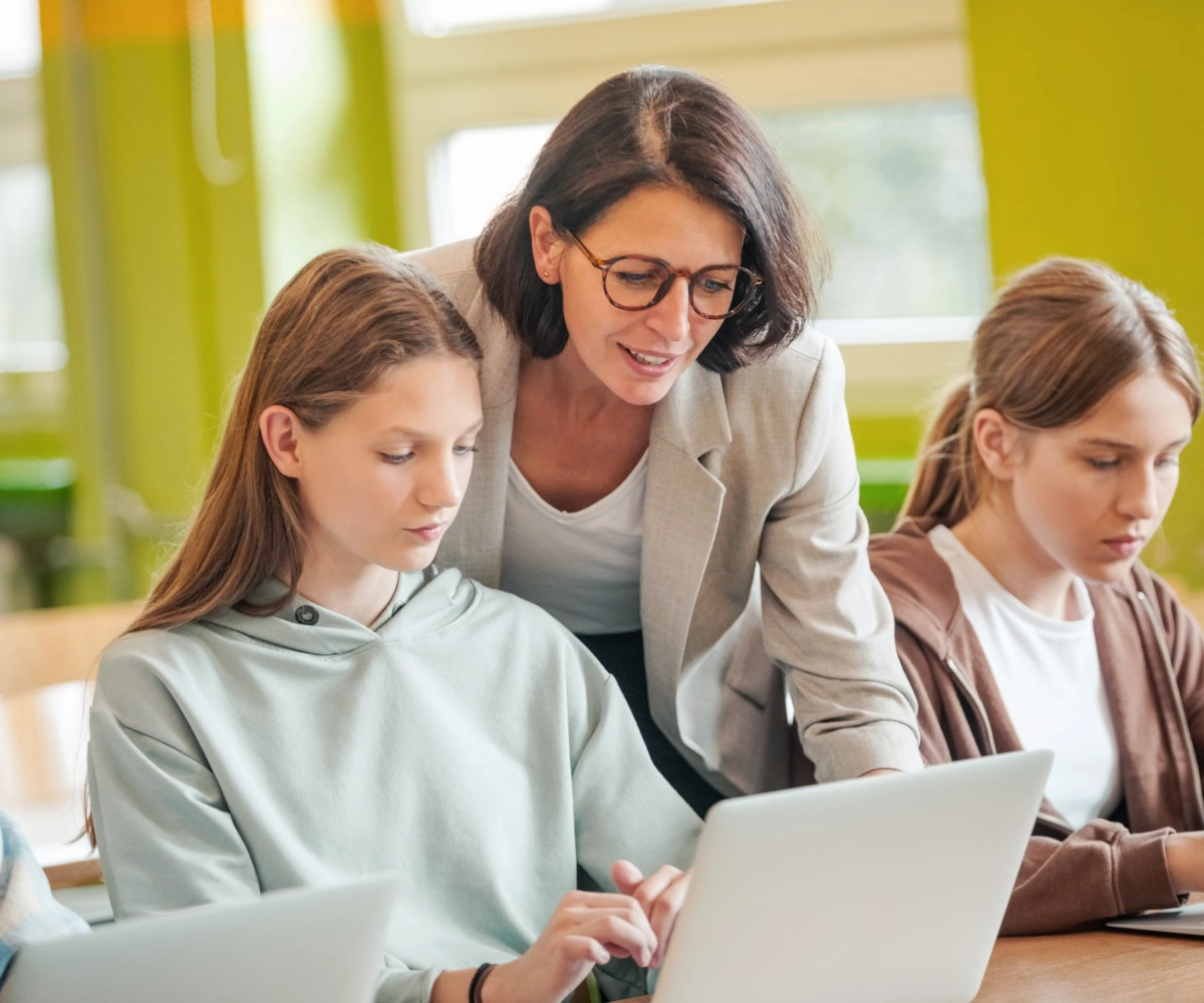 To tenåringsjenter sitter med en laptop foran seg, mens en voksen kvinne med svarte briller bøyer seg over den ene jenta og ser på skjermen hennes. I bakgrunnen står en gul bygning med store vinduer. Foto.