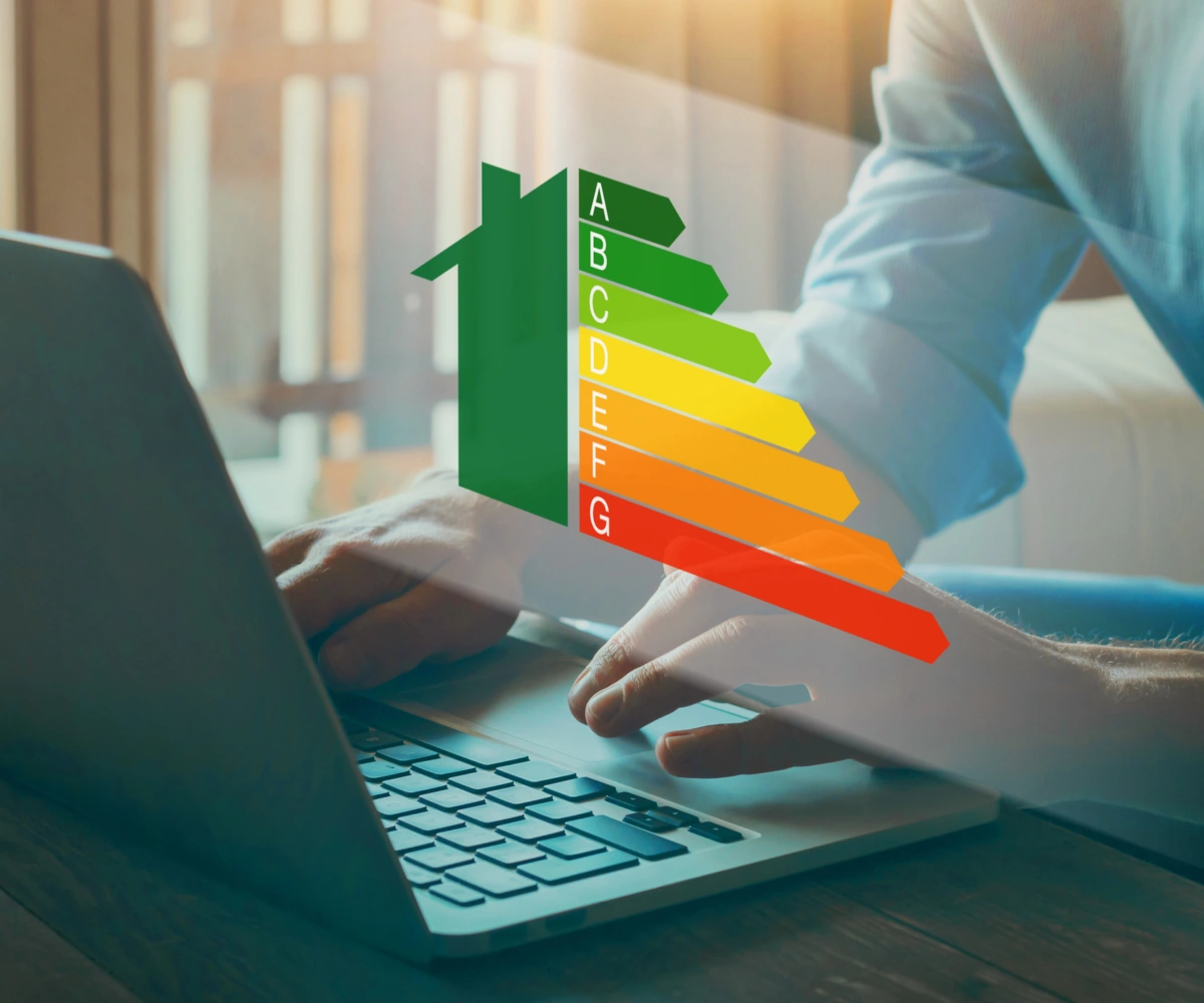 Nærbilde av baksiden av en laptop med et par hender over tastaturet. Over hendene vises en illustrasjon av et halvt grønt hus med energimerking fra A til G i en fargeskala fra grønt til rødt. Foto og illustrasjon.
