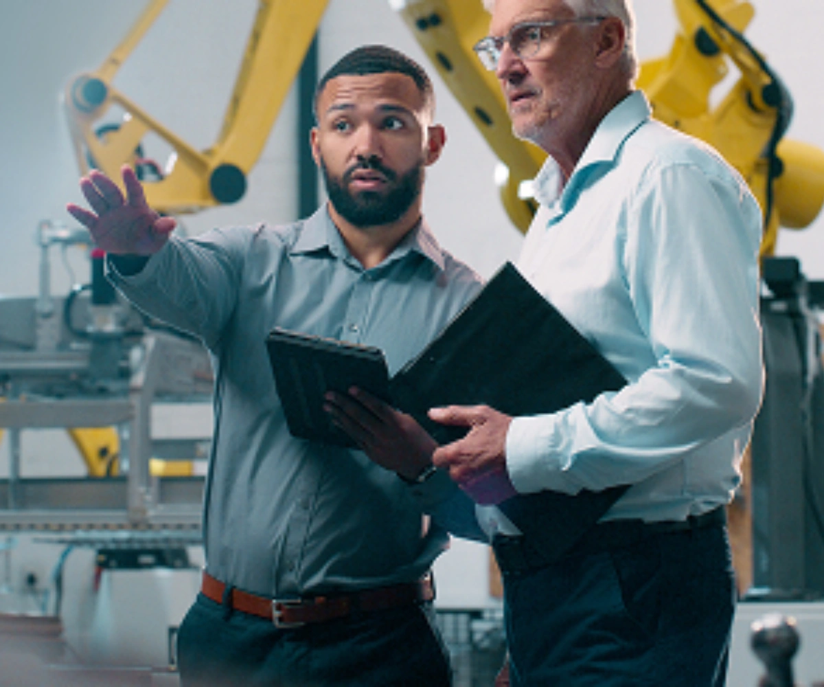 To menn i skjorter står i en fabrikk. Den ene peker utover fabrikken, mens den andre følger blikket i samme retning. I bakgrunnen sees maskiner og en gul kran. Bilde.