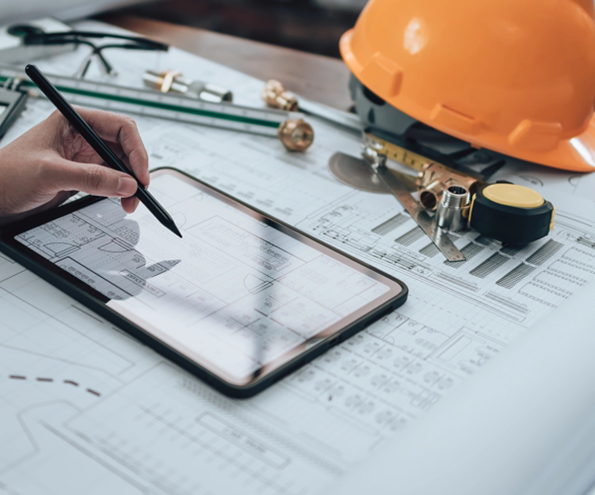 Nærbilde av en pult med arkitekttegninger, en gul vernehjelm, linjaler og diverse arbeidsverktøy. En hånd med en svart penn peker på en nettbrett som ligger oppå tegningene. Bilde.