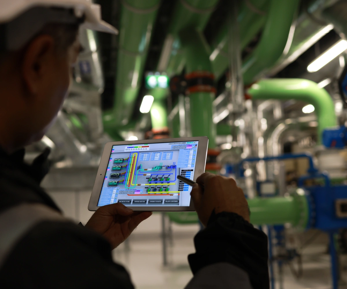 En industriarbeider iført vernehjelm ser på en tablet med målinger. I bakgrunnen er grønne og hvite rør synlige fra taket og veggene. Foto.