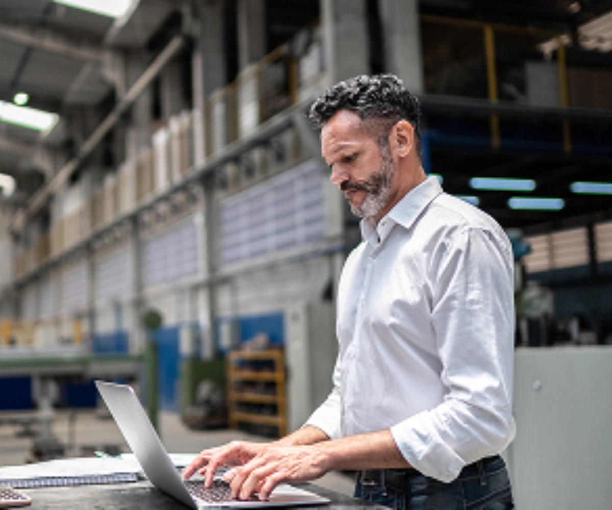 En mann i lys skjorte jobber stående på en laptop. I bakgrunnen sees et industribygg. Bilde.