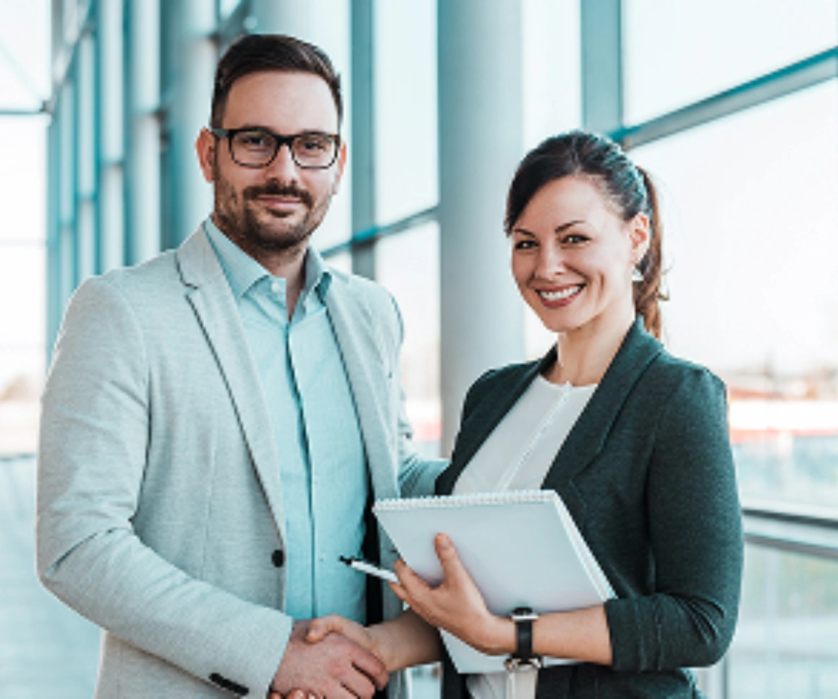 En mann og en kvinne i skjortebluser smiler. I bakgrunnen sees et glassbygg. Bilde.