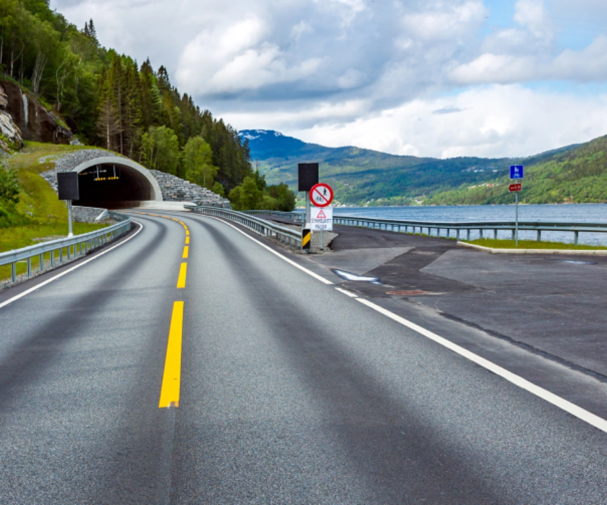 En motorvei leder inn i en tunnel i forgrunnen. Over tunnelen vokser grønne trær, og til høyre for den ligger et vann omgitt av fjellkjeder. Foto.