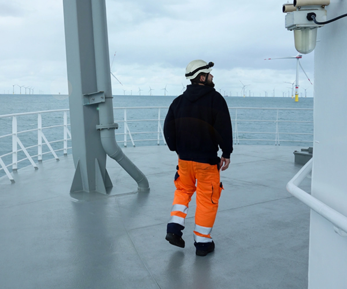 En mann iført oransje arbeidsbukse og hvit vernehjelm går på en plattform med hvite rekkverk. Bak rekkverket strekker havet seg ut, med mange vindmøller synlige i det fjerne. Foto.