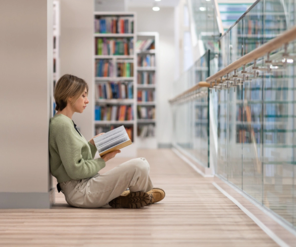 En kvinne sitter med beina i kryss på gulvet, lent inntil en søyle nær et glassrekkverk, mens hun blar i en bok. I bakgrunnen står store bokhyller fylt med mange bøker. Foto.