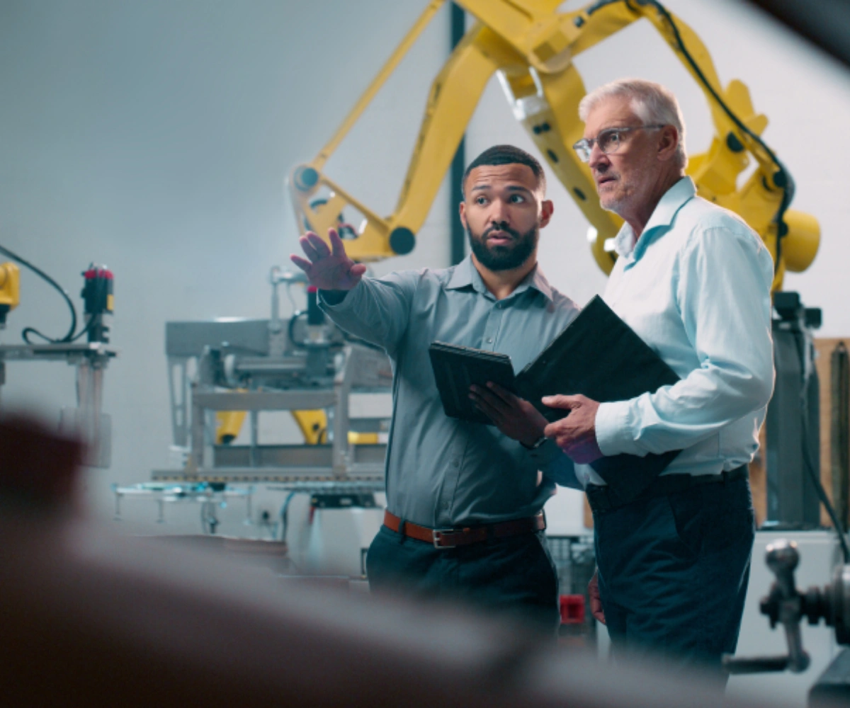 To menn i skjorter står i en fabrikk. Den ene peker utover fabrikken, mens den andre følger blikket i samme retning. I bakgrunnen sees maskiner og en gul kran. Foto.