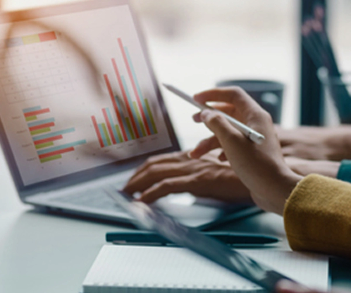 Nærbilde av en laptopskjerm som viser grafer, med et par hender på tastaturet. En annen hånd holder en penn og peker mot grafene på skjermen.