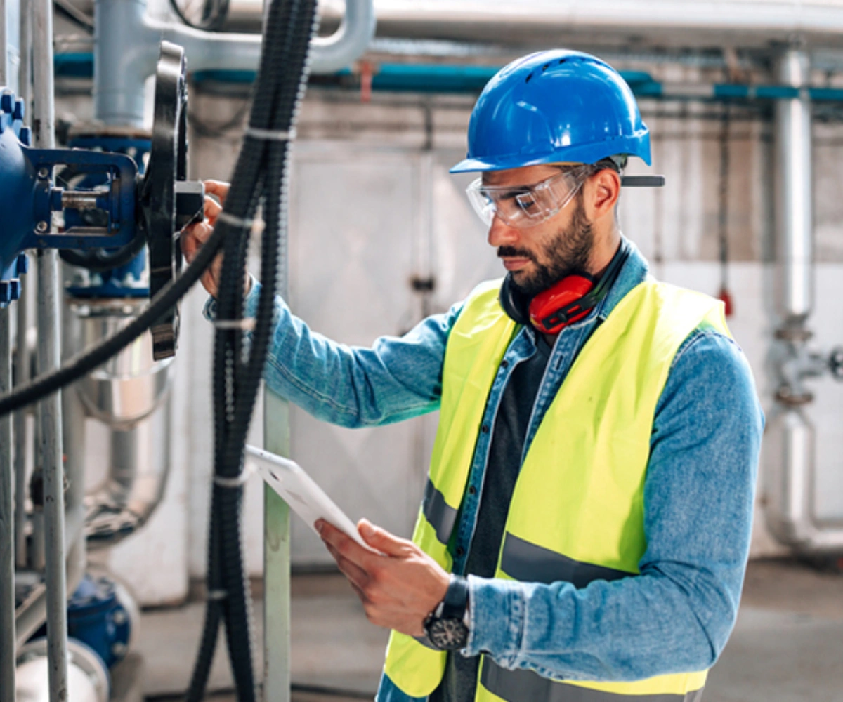 En person iført gul vest, vernebriller, hjelm og hørselsvern rundt halsen studerer en rapport. I bakgrunnen er en betongvegg med synlige ledninger. Foto.