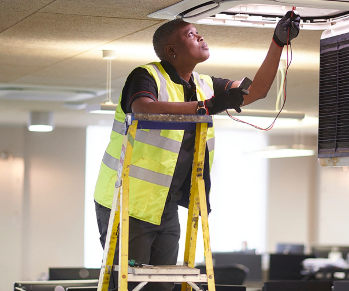 En mann i gul vest står på en gul stige og inspiserer elektriske ledninger i takhimlingen. Foto.