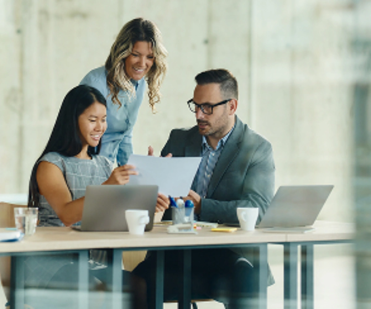 Tre personer smiler og diskuterer ved et bord. Den ene sitter og holder en gjenstand, den andre sitter og peker, mens den tredje står lett bøyd over dem i midten. Foto.