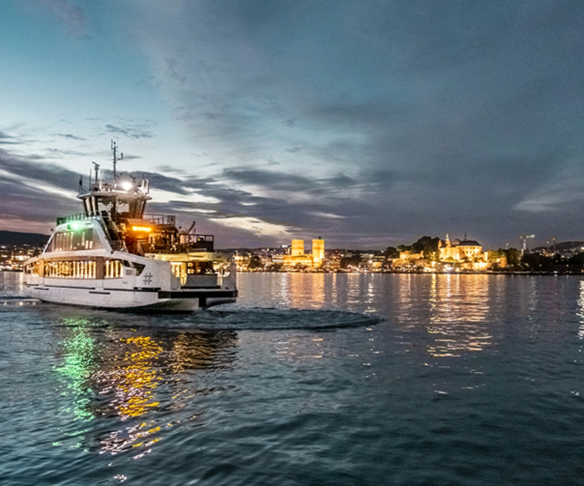 En opplyst båt på det mørke vannet like før solnedgang, med en opplyst by i bakgrunnen. Foto.