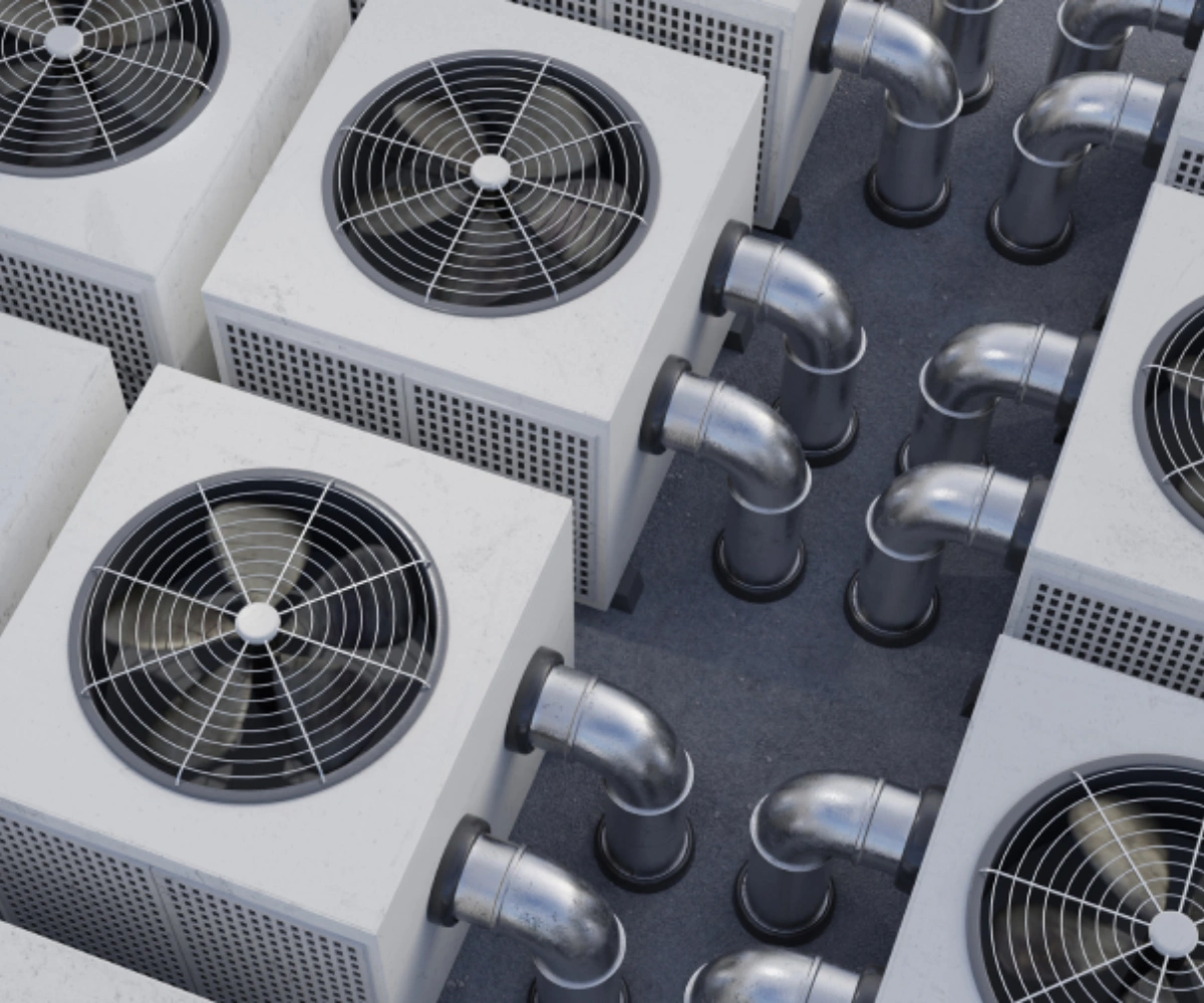 Flere store HVAC-enheter (varme-, ventilasjons- og klimaanlegg) med metalliske vifter og rør ligger plassert i et industrielt utendørsmiljø. Sølvfargede rør strekker seg ut fra enhetene og går i ulike retninger. Foto.