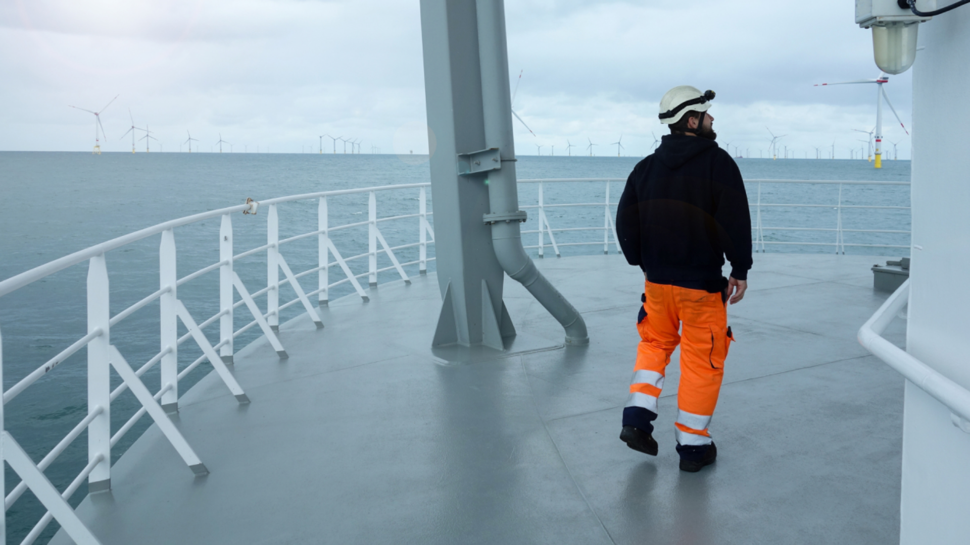 En mann iført oransje arbeidsbukse og hvit vernehjelm går på en plattform med hvite rekkverk. Bak rekkverket strekker havet seg ut, med mange vindmøller synlige i det fjerne. Foto.